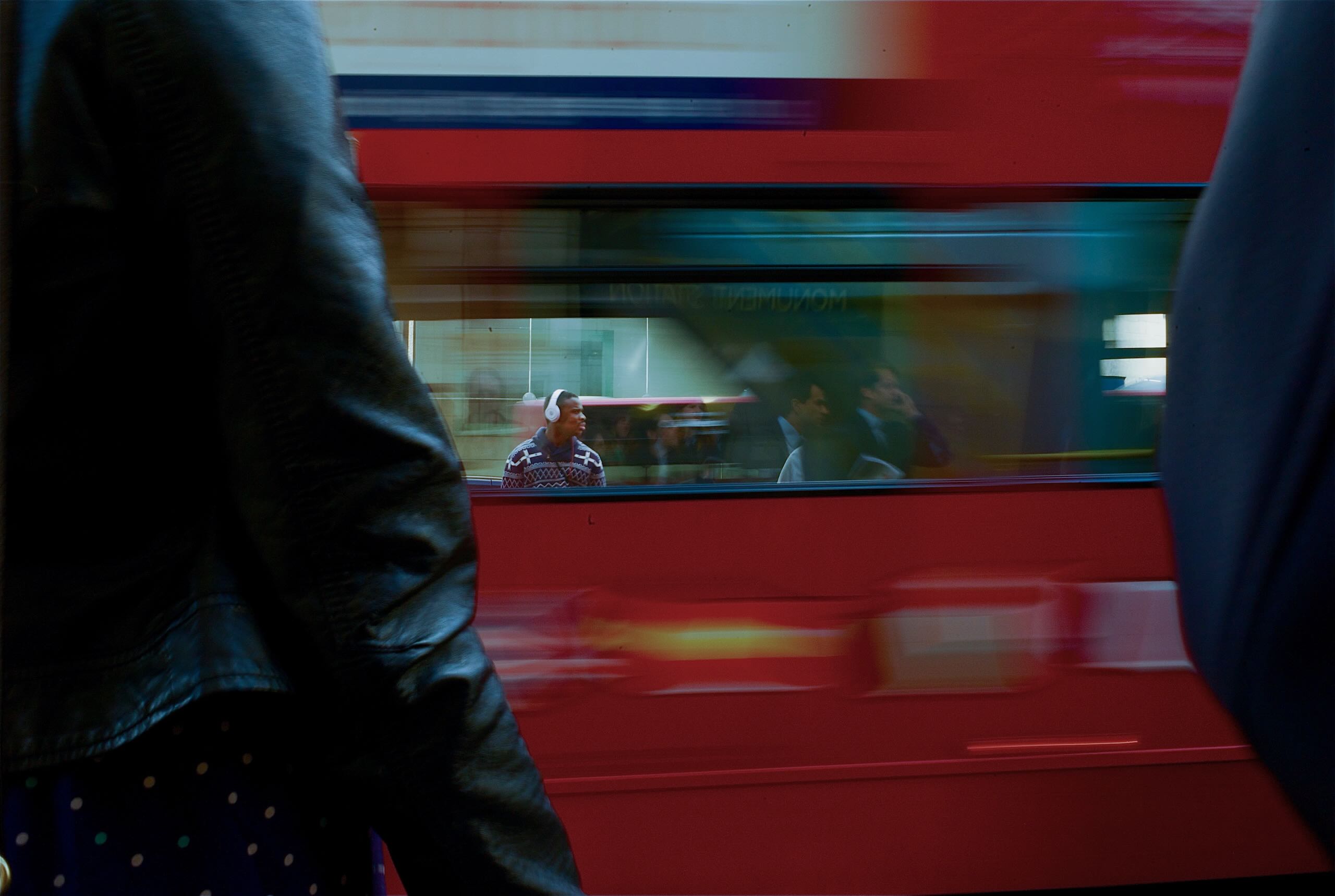 Street photograph in London shot through a passing red bus, showing a casually dressed man and businessmen in suits through the bus windows